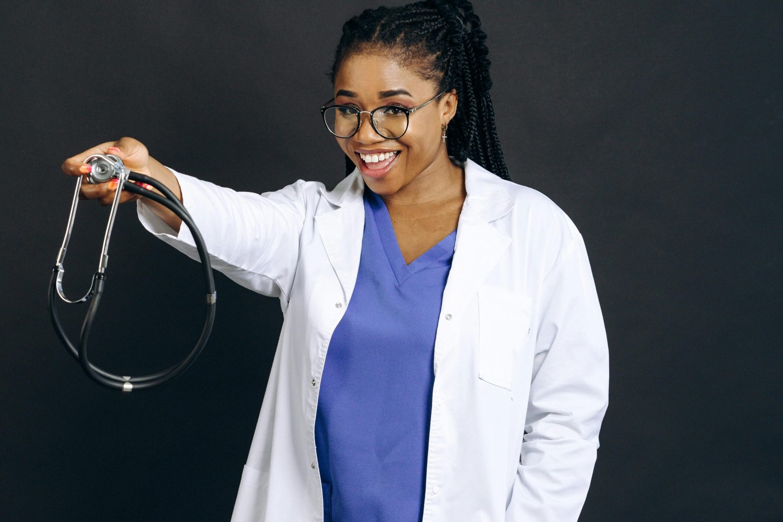 Confident female doctor in white coat smiling and holding a stethoscope against a black background.