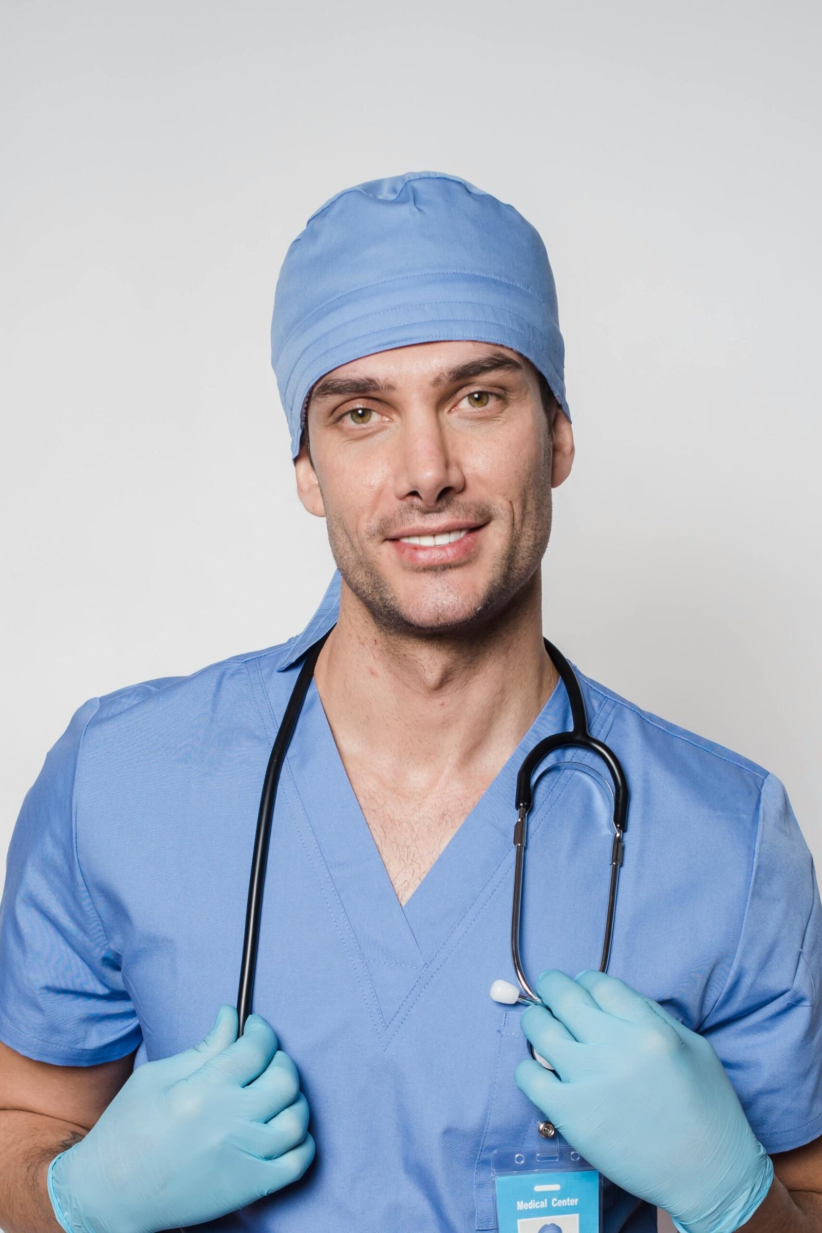 Positive man doctor in blue uniform and protective gloves standing with stethoscope and looking at camera against light background